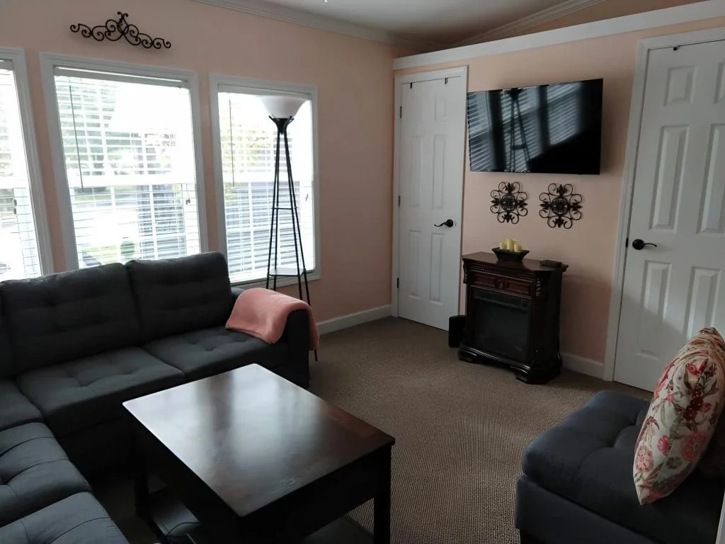 Comfortable family room featuring large windows, an electric fireplace, and wall-mounted TV inside the Crimson Poppy tiny home.